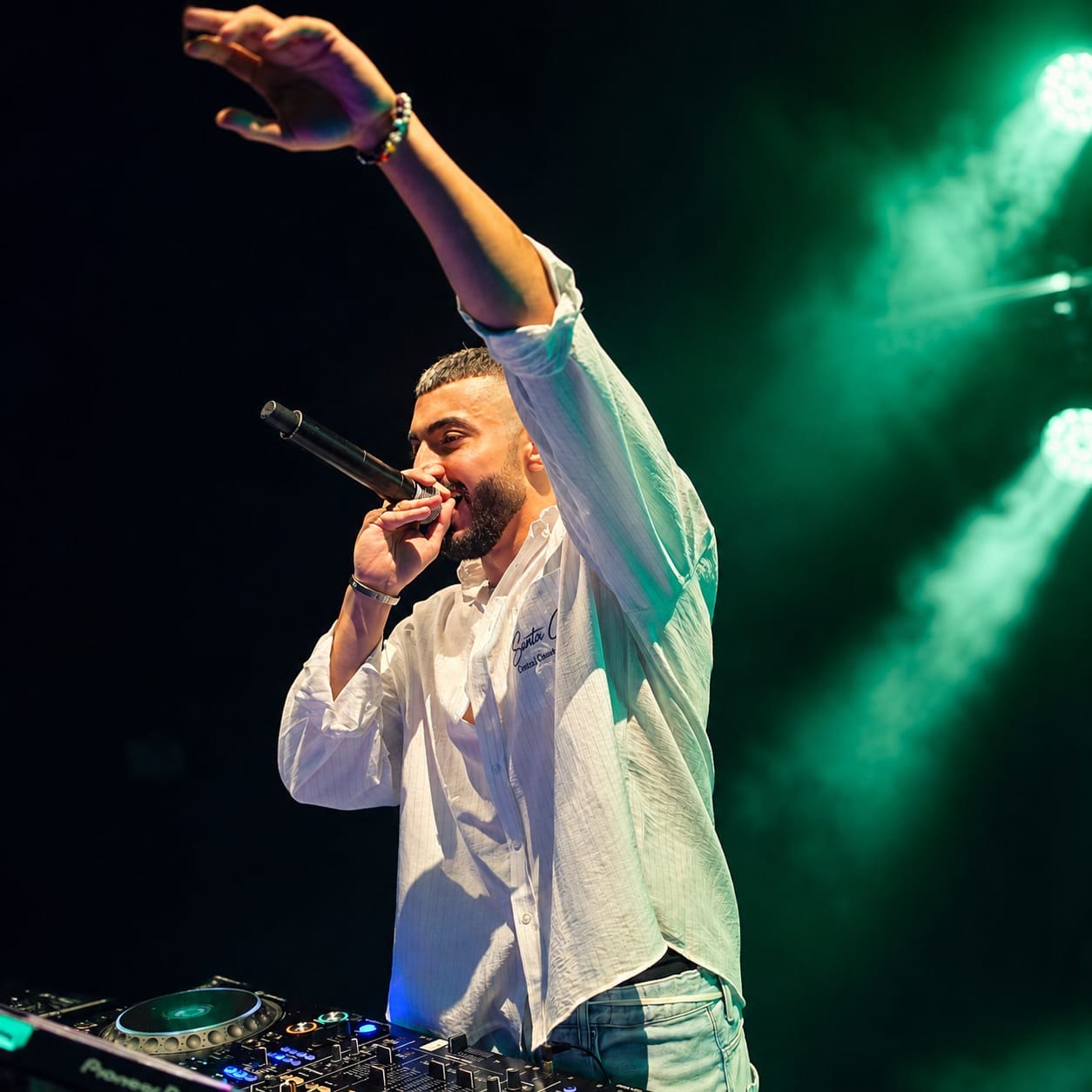 Live performer singing into a microphone with one arm raised above the DJ booth